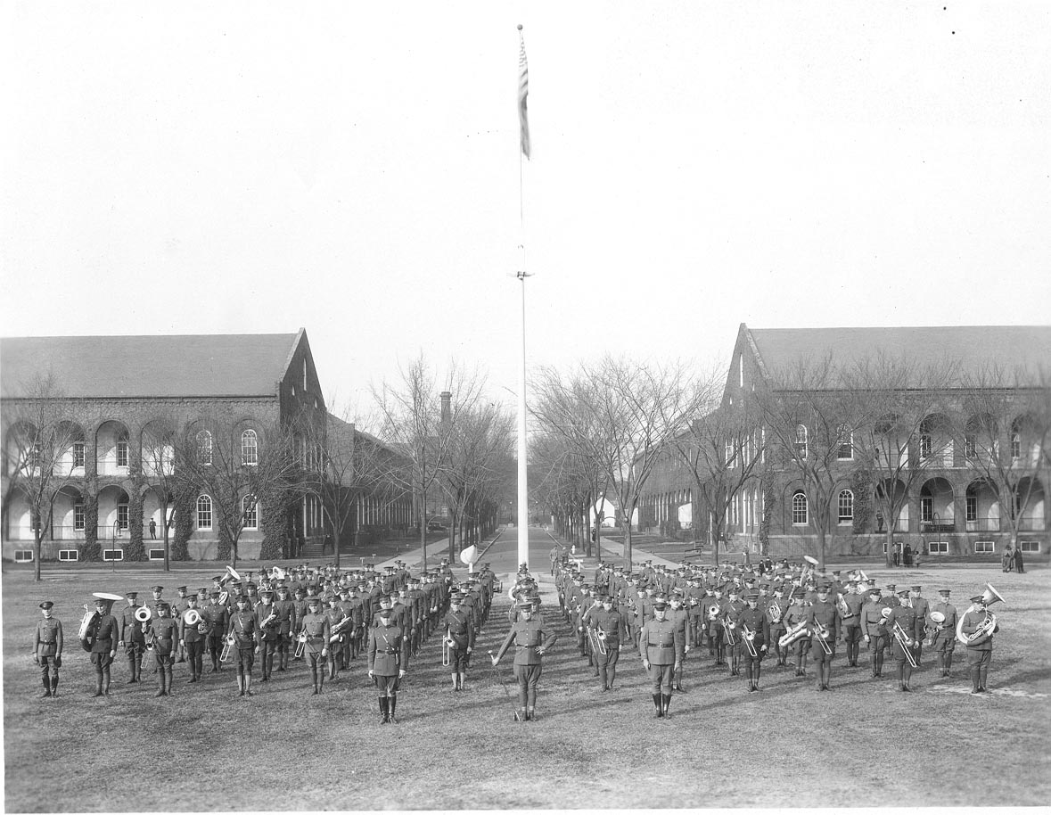 Black and white historic image of a large military band standing in rows/formation on a grass field in front of a white flagpole and two 2-story brick buildings.